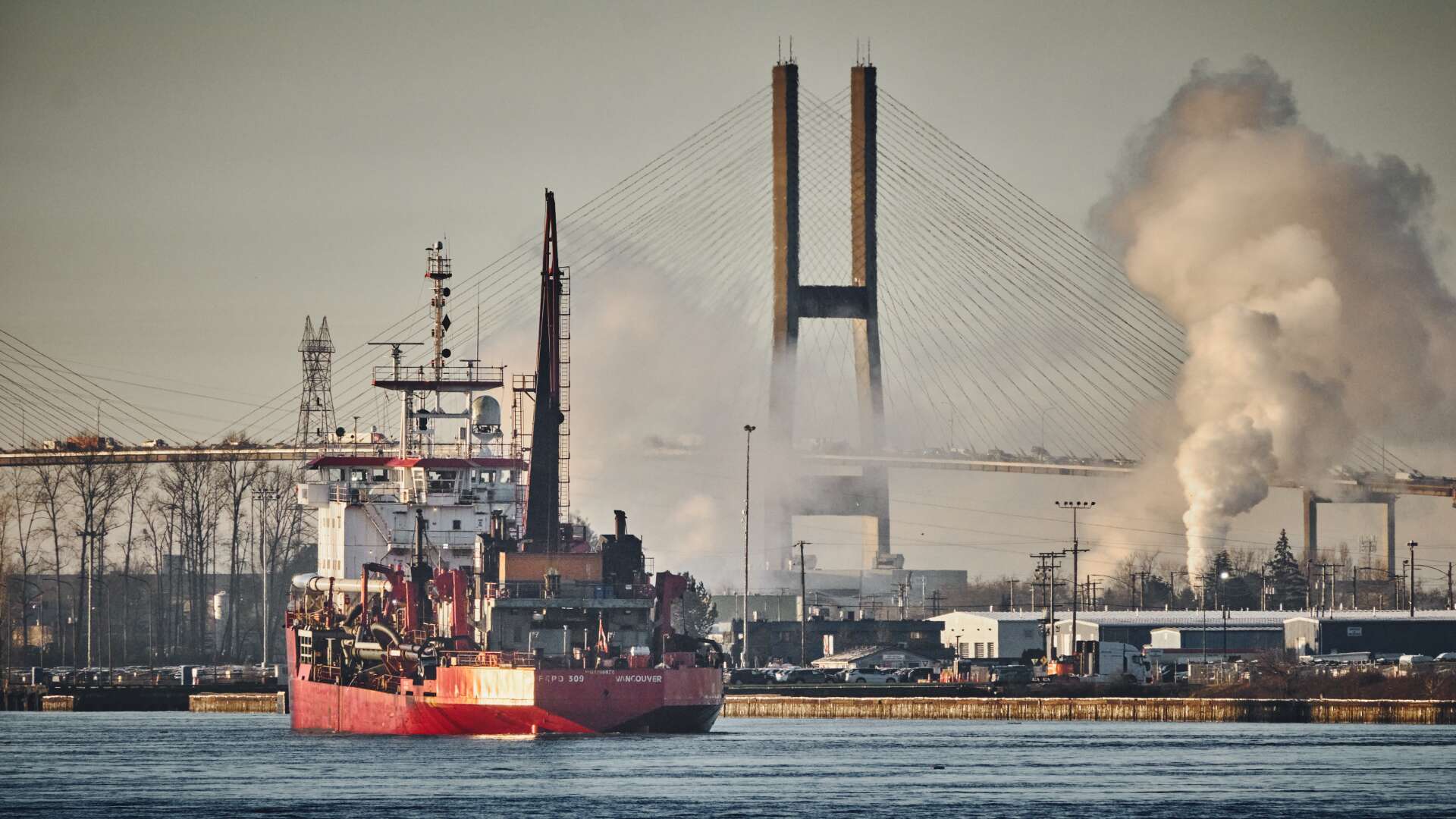 A photo depicting a boat on the Fraser River with a backdrop of the Alex Fraser bridge and industrial buildings.